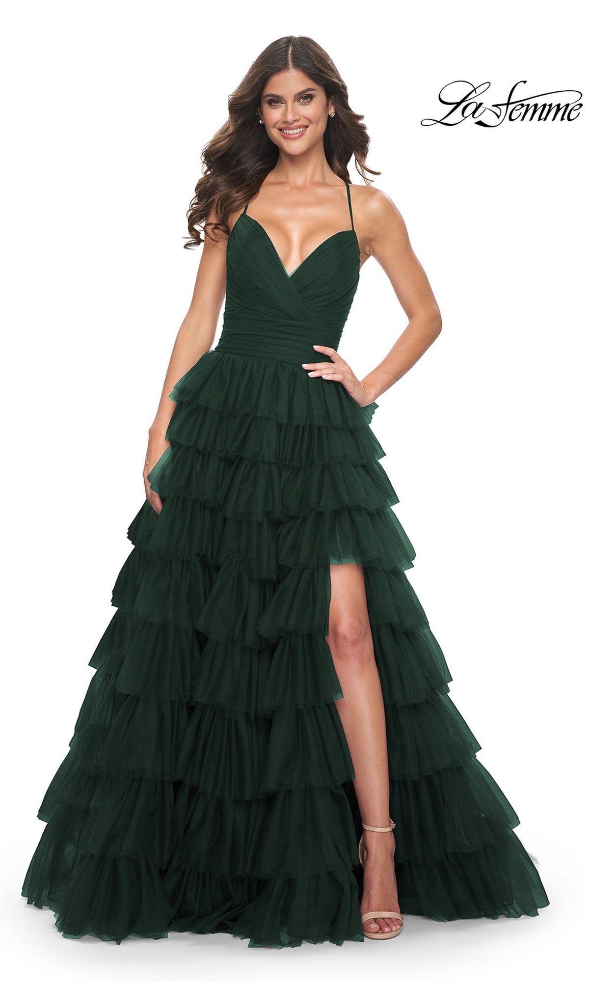 A woman wearing the La Femme Lace-Up Long Tiered Prom Dress 32086 in Dark Emerald, featuring ruffled details, a deep V-neckline, spaghetti straps, and a high slit, stands against a white background.